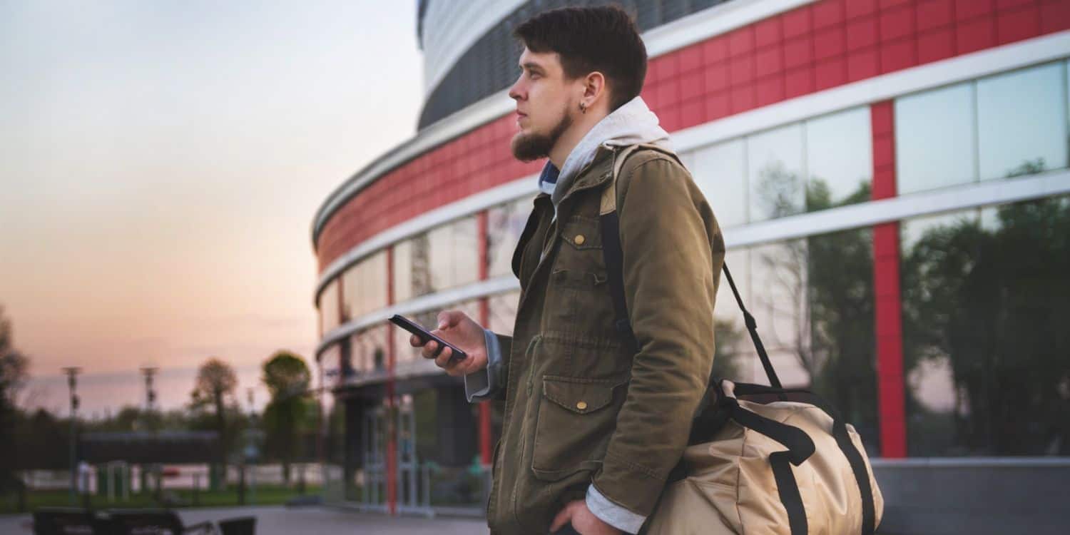 Férfi áll egy épület előtt kezében a telefonja, vállán pedig egy sporttáska.
